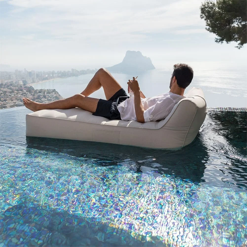 A man relaxing on a white sofa at the edge of an infinity pool overlooking a coastal city and a mountain in the distance.
