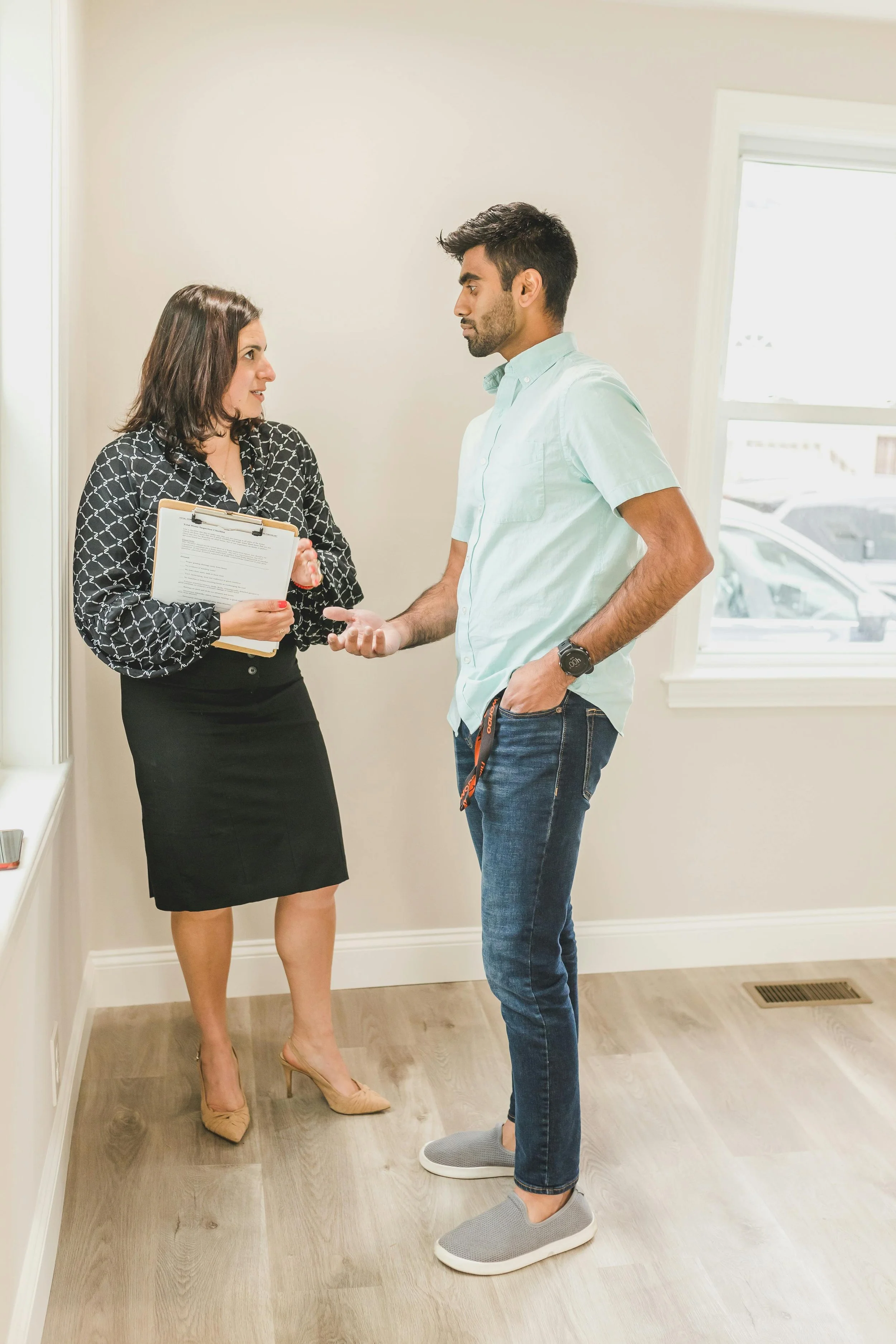 A woman holding documents and speaking with a man inside a room with a window, white walls, and wooden flooring.