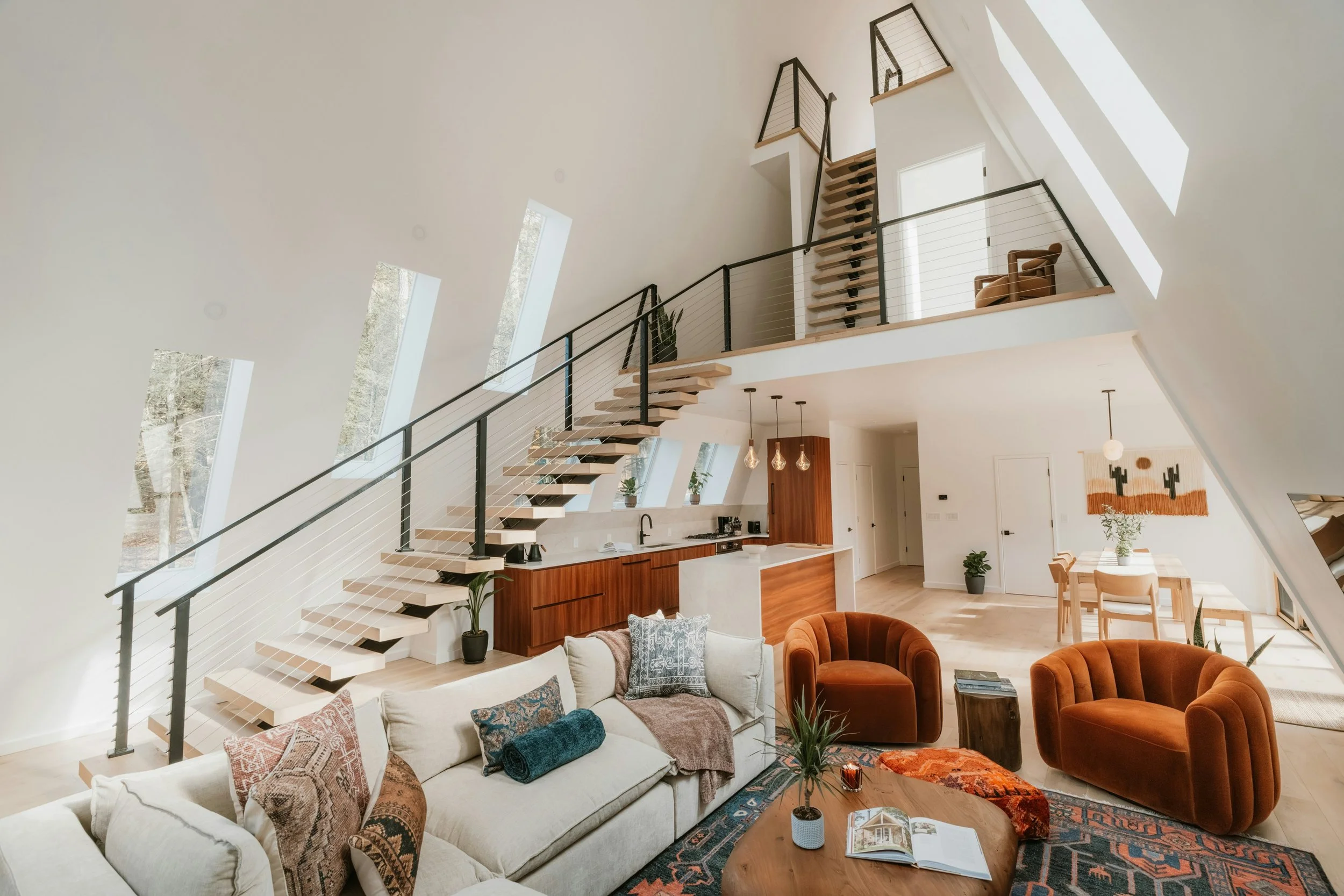 Interior view of a modern, open-plan living space with a high, sloped ceiling and large windows. There is a kitchen with wooden cabinets and a white island, a cozy seating area with a light-colored sofa, orange armchairs, a round wooden coffee table, and a dining area with a table and chairs. The space features natural light, potted plants, and contemporary decor.
