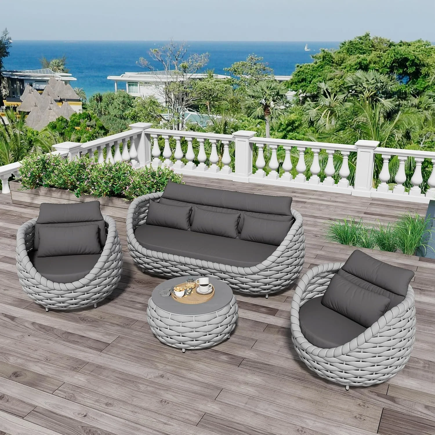 A modern outdoor patio with a view of the ocean, featuring wicker furniture with gray cushions, a small matching table with coffee cups and snacks, surrounded by lush greenery and flowers.
