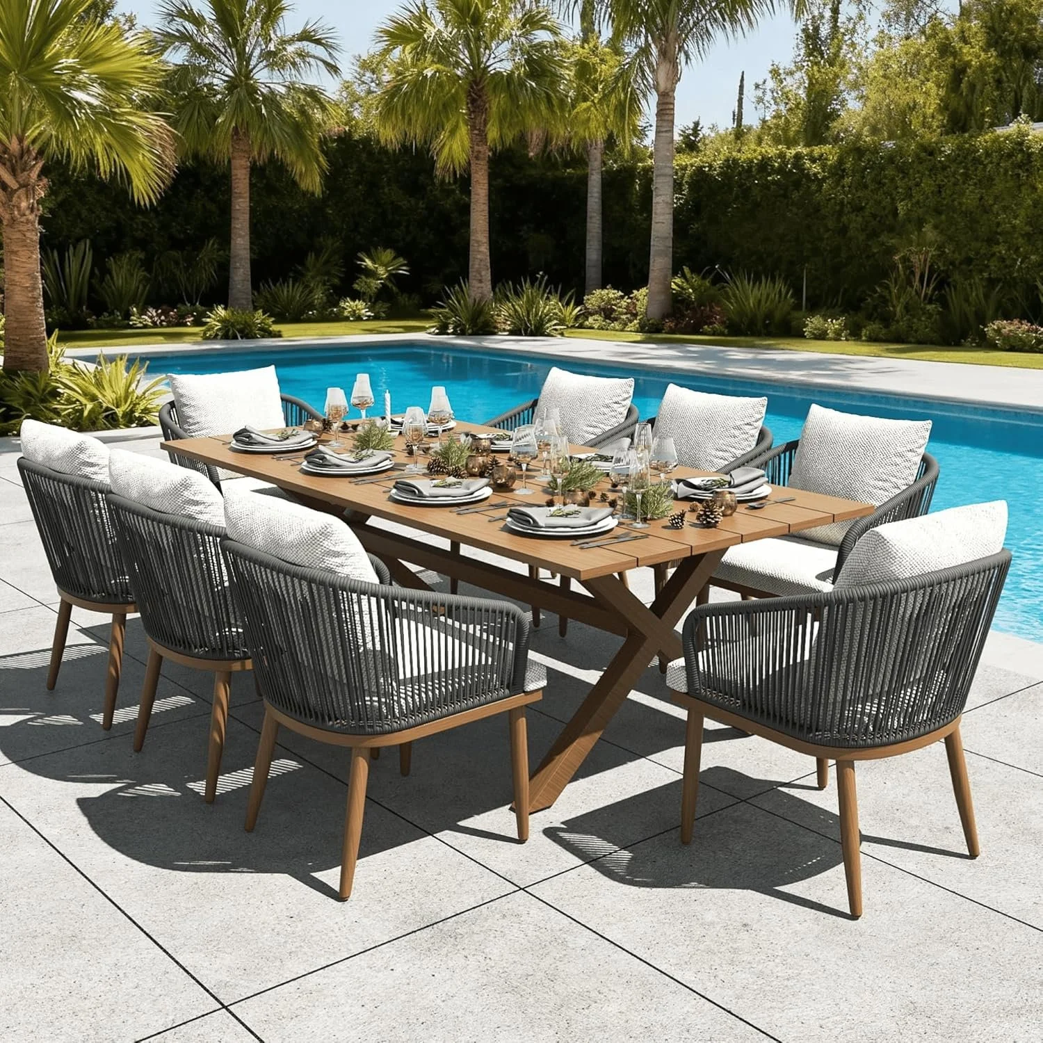 Outdoor dining table set by a pool with palm trees, shrubs, and clear sky in the background.