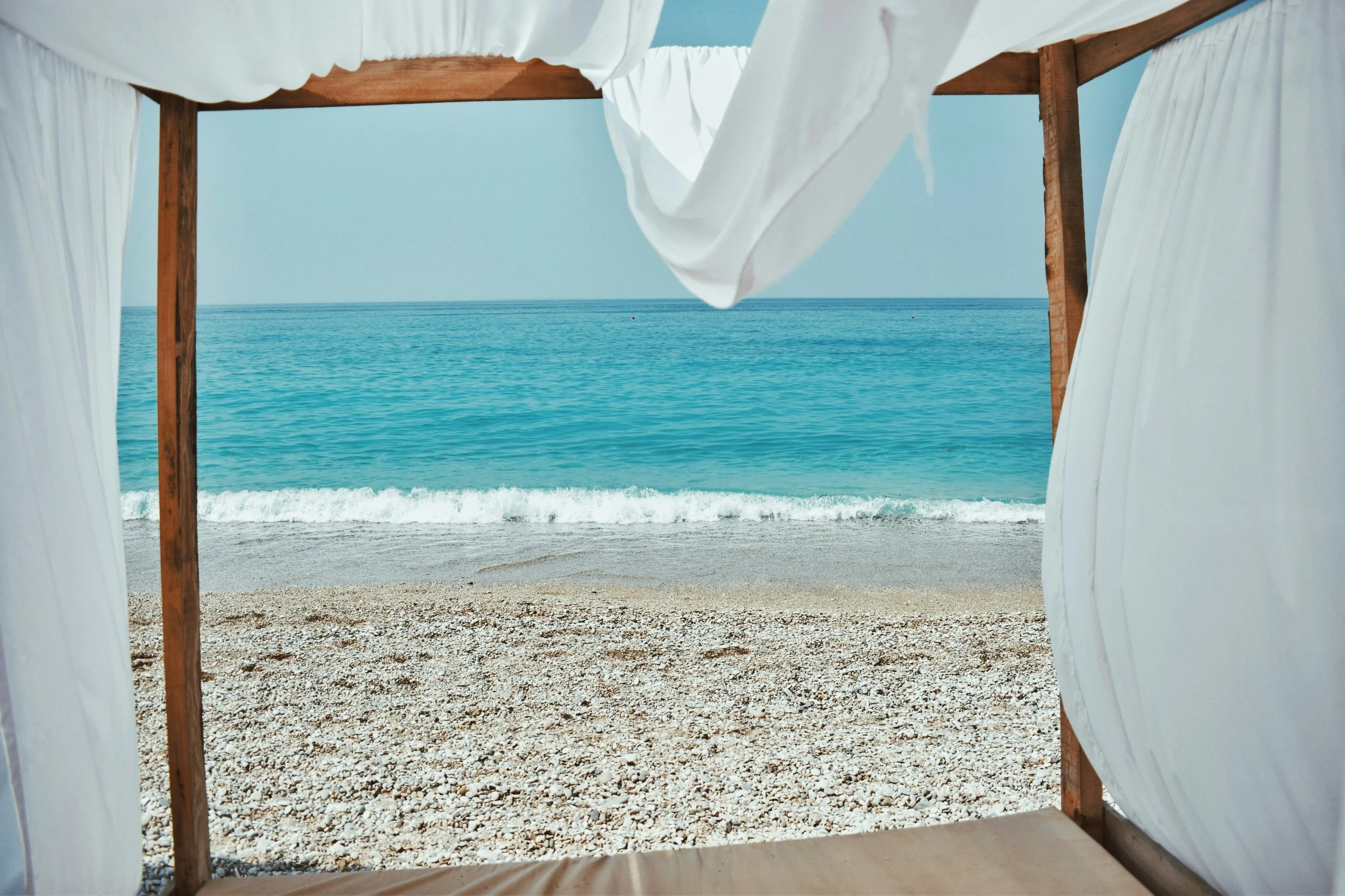 View of a beach with pebbles, gentle waves, and turquoise water seen from a wooden cabana with white curtains.