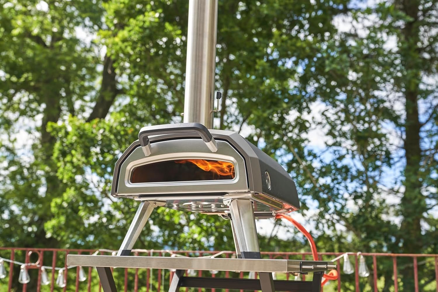 Outdoor pizza oven with a small flame burning inside, set on a metal stand with trees and string lights in the background.