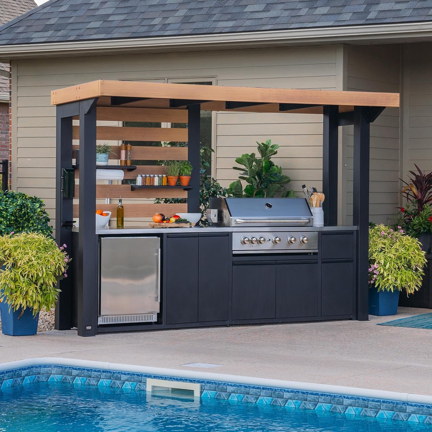 Outdoor kitchen area with a built-in grill, stainless steel mini refrigerator, black cabinetry, wooden countertop, and a wooden roof extension, surrounded by potted plants, near a swimming pool.
