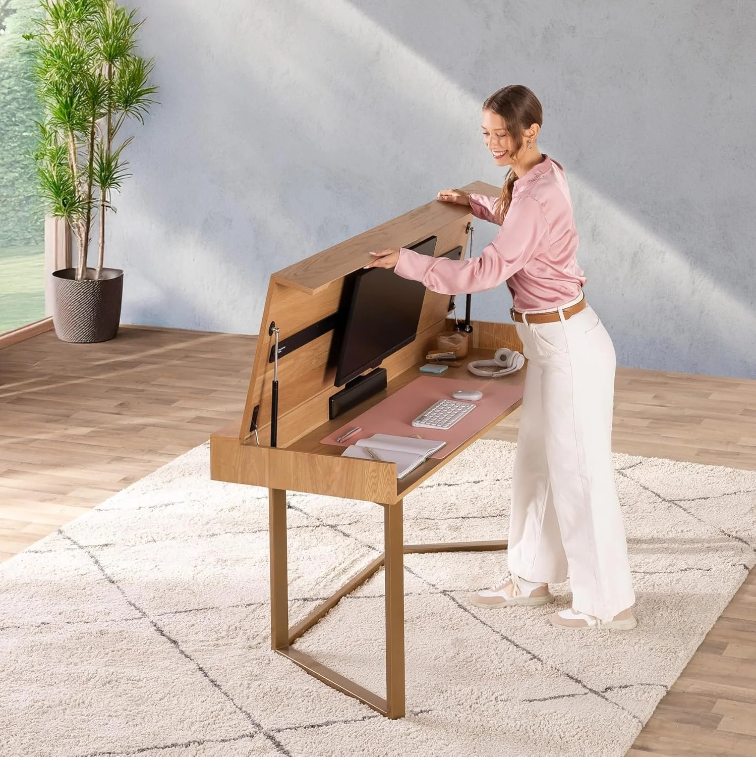 A woman stands at a modern wooden desk with a tilted top, working with a computer setup in a bright room with wooden floors, a white rug, and a potted plant in the background.