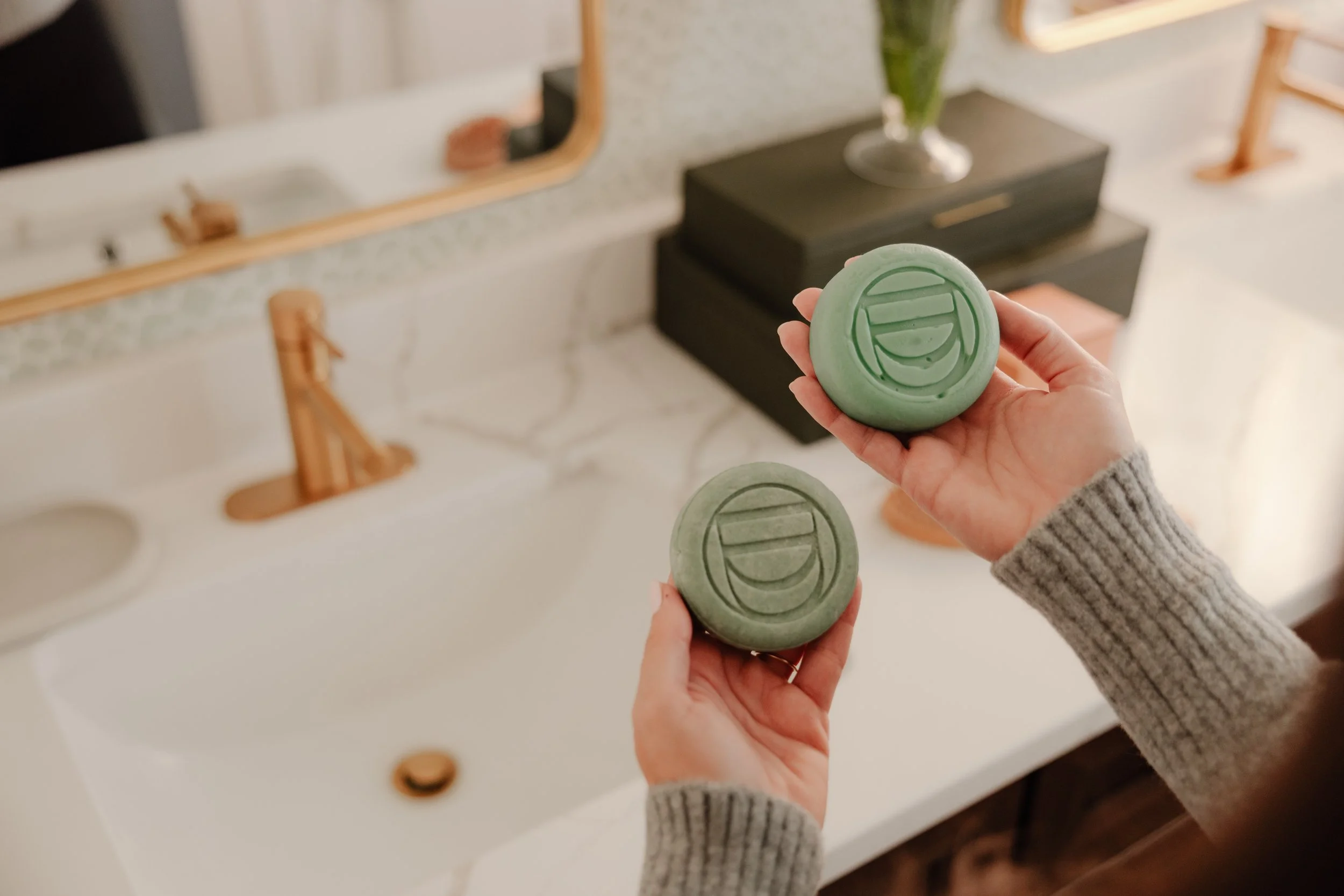 Two hands holding round, green soap bars with circular designs in front of a white bathroom sink and mirror.
