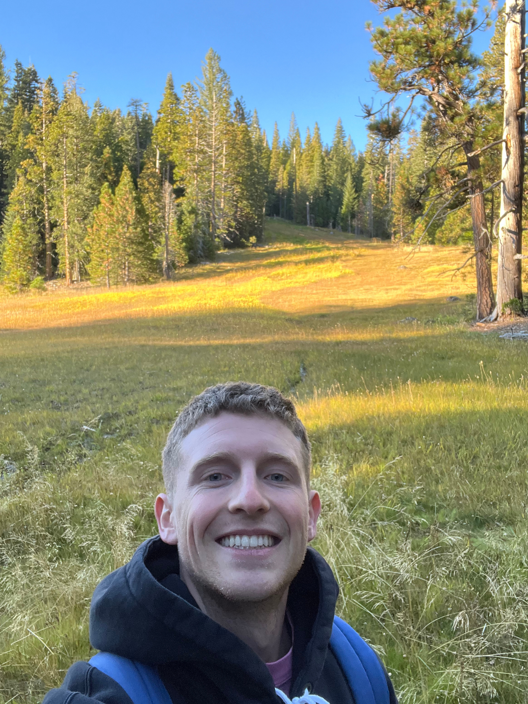 A man with short light brown hair smiling outdoors in a grassy meadow, with a backdrop of green pine trees and a clear blue sky, wearing a black hoodie and carrying a blue backpack.