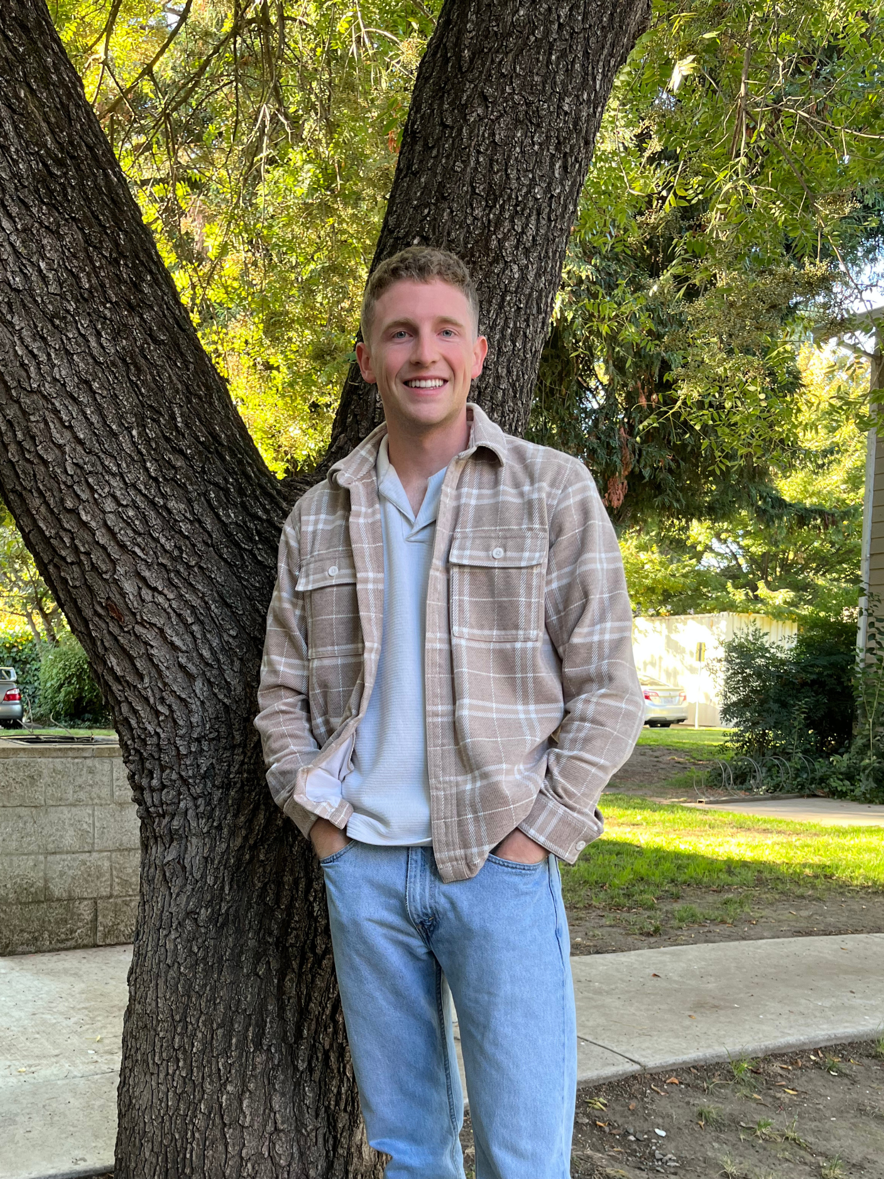 A young man with short light brown hair smiling, wearing a beige plaid shirt over a white t-shirt and light blue jeans, standing outdoors next to a large tree in a park or yard with green foliage and parked cars in the background.
