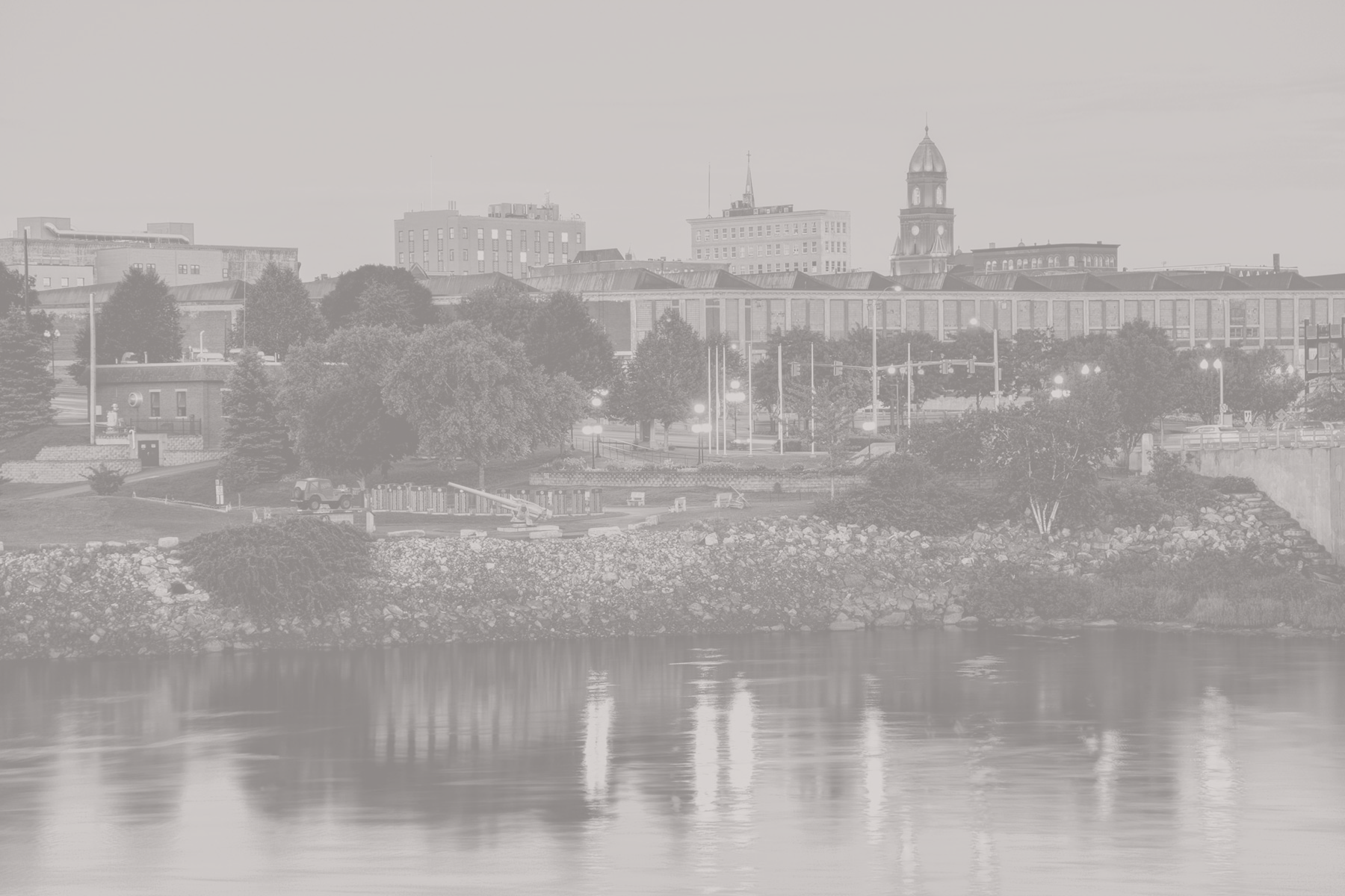 A grayscale cityscape of Lewiston, Maine, showing buildings, trees, a waterfront, and a clock tower in the distance.