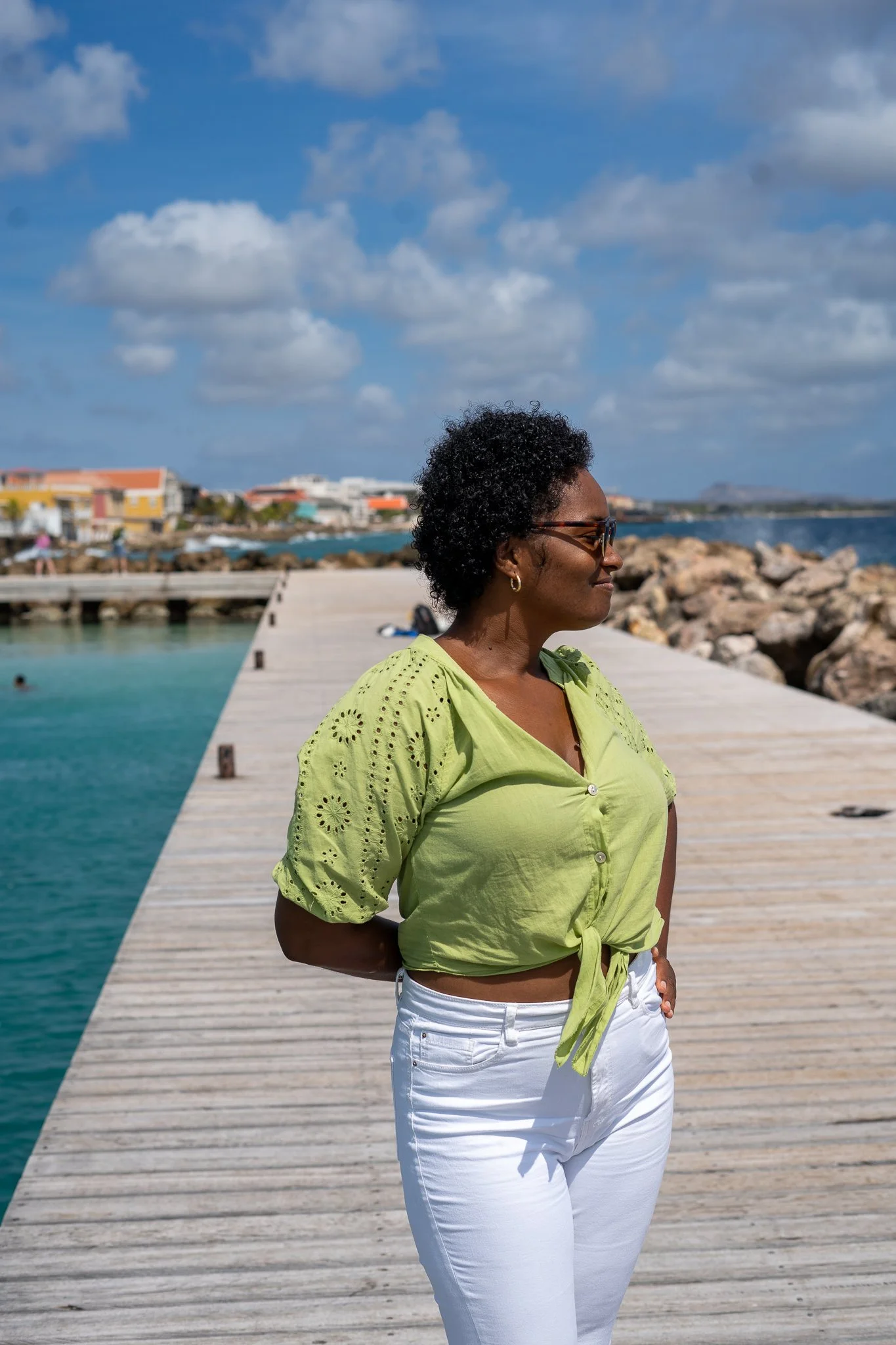 A woman standing on a pier by the water, wearing a light green blouse and white pants, with short curly black hair and sunglasses, smiling as she looks to her right, with a background of buildings, rocks, and a cloudy sky.