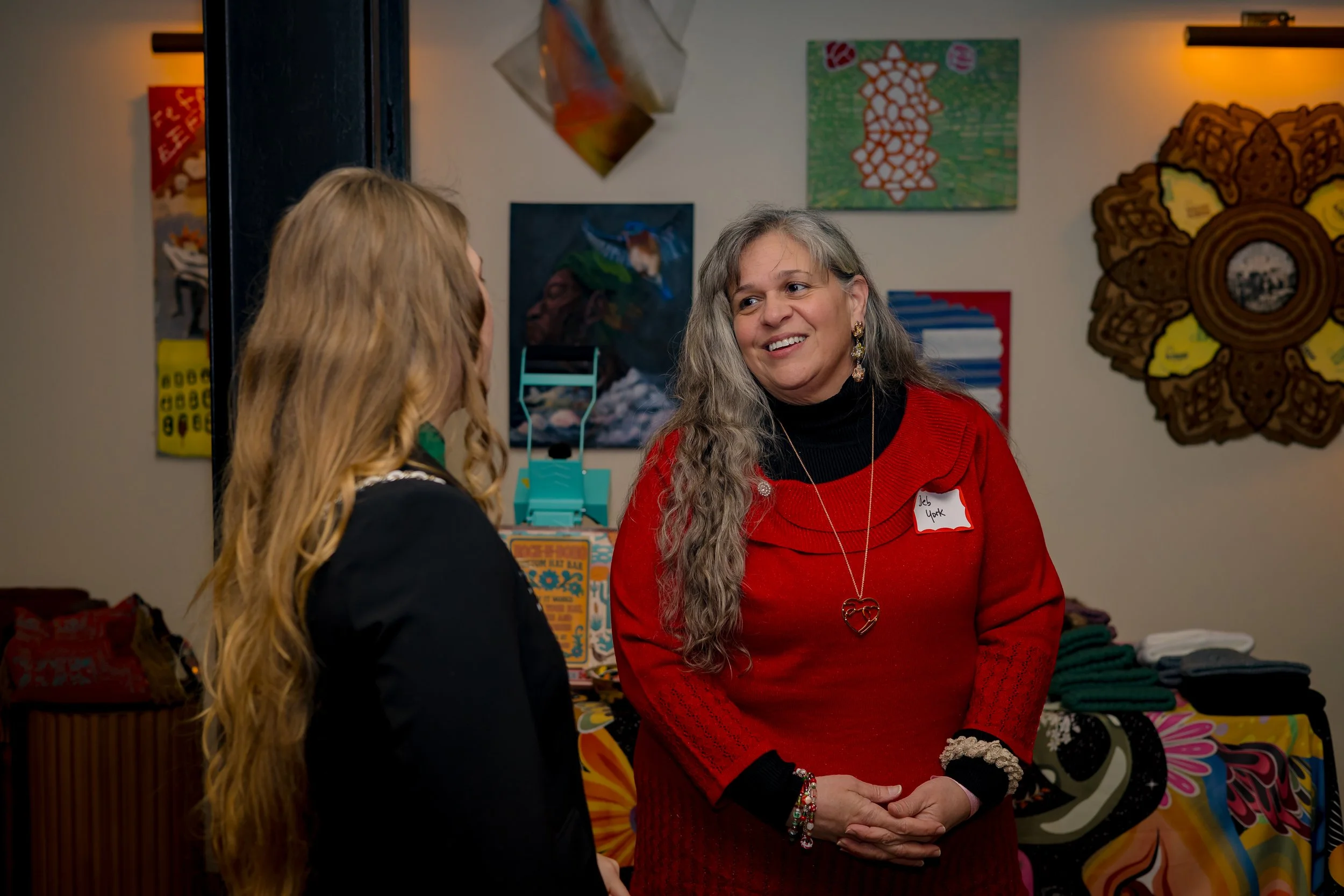 Two women having a conversation at an indoor event, with colorful artwork and decorations in the background.