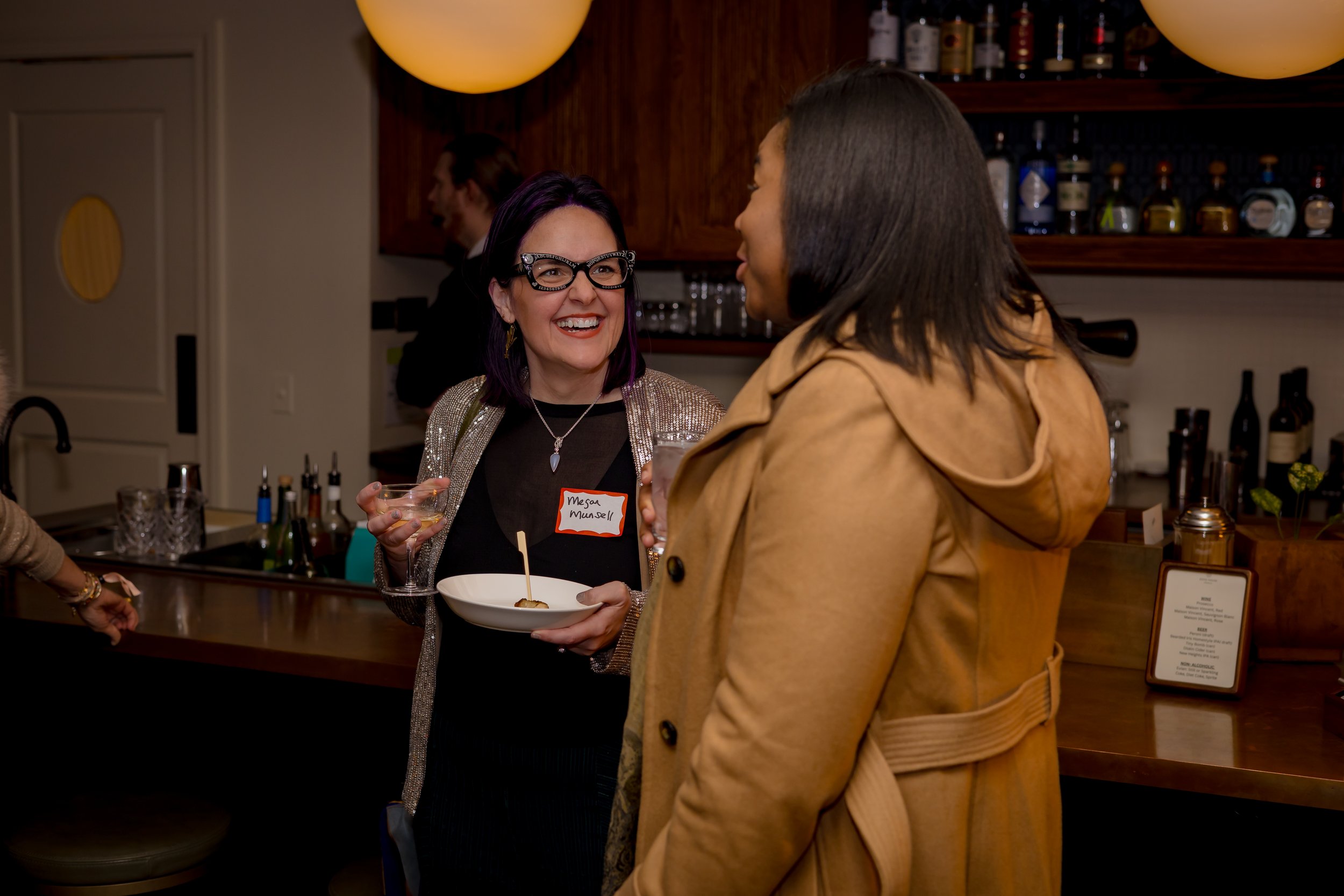 Two women talking and smiling at a bar, one holding a drink and a plate with a skewer, the other wearing a tan coat. Background includes bar shelves with bottles and a menu stand.