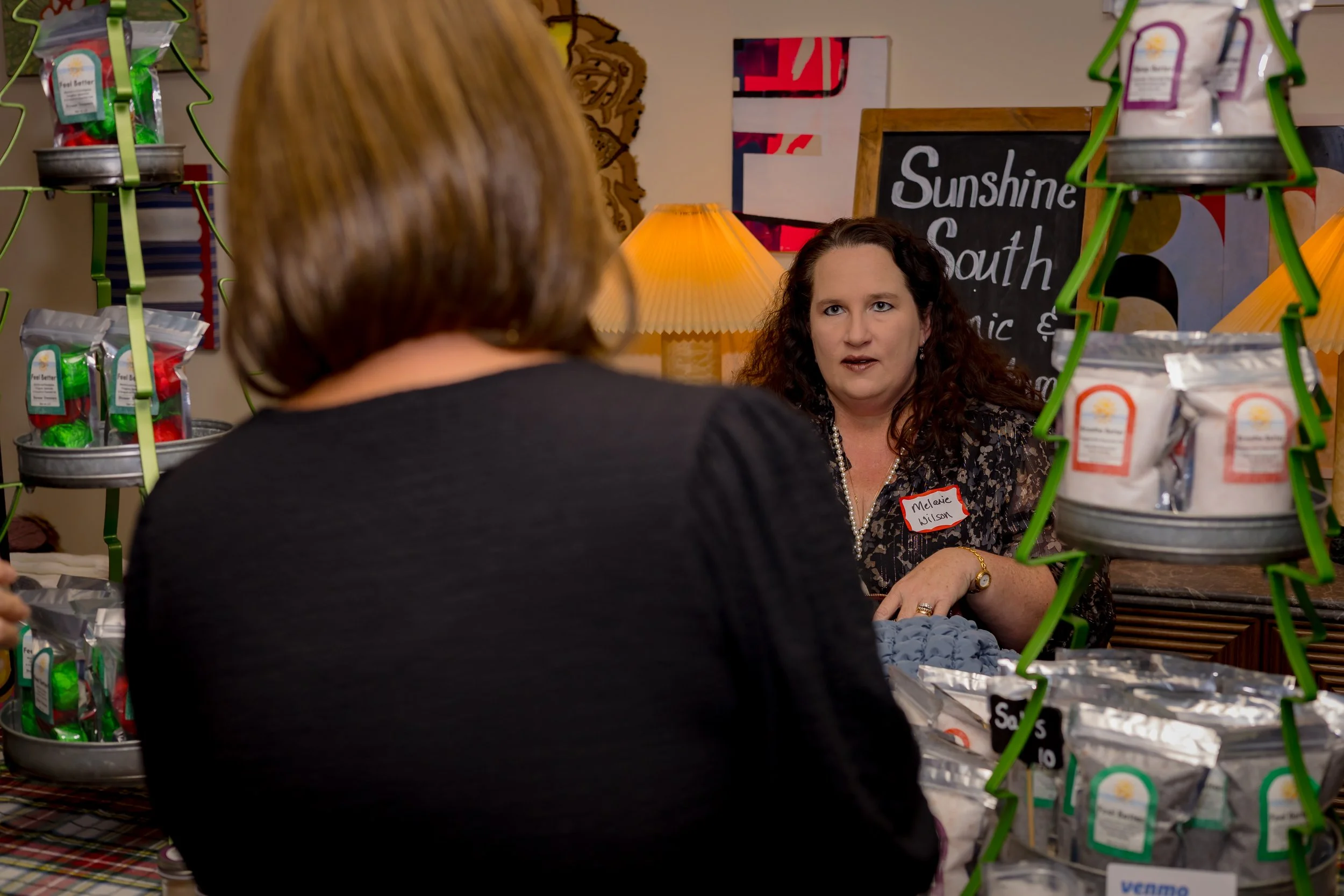 A woman with curly dark hair and wearing a black floral blouse talks to another woman whose back is to the camera, at a colorful market stall with bags of products and a chalkboard sign.