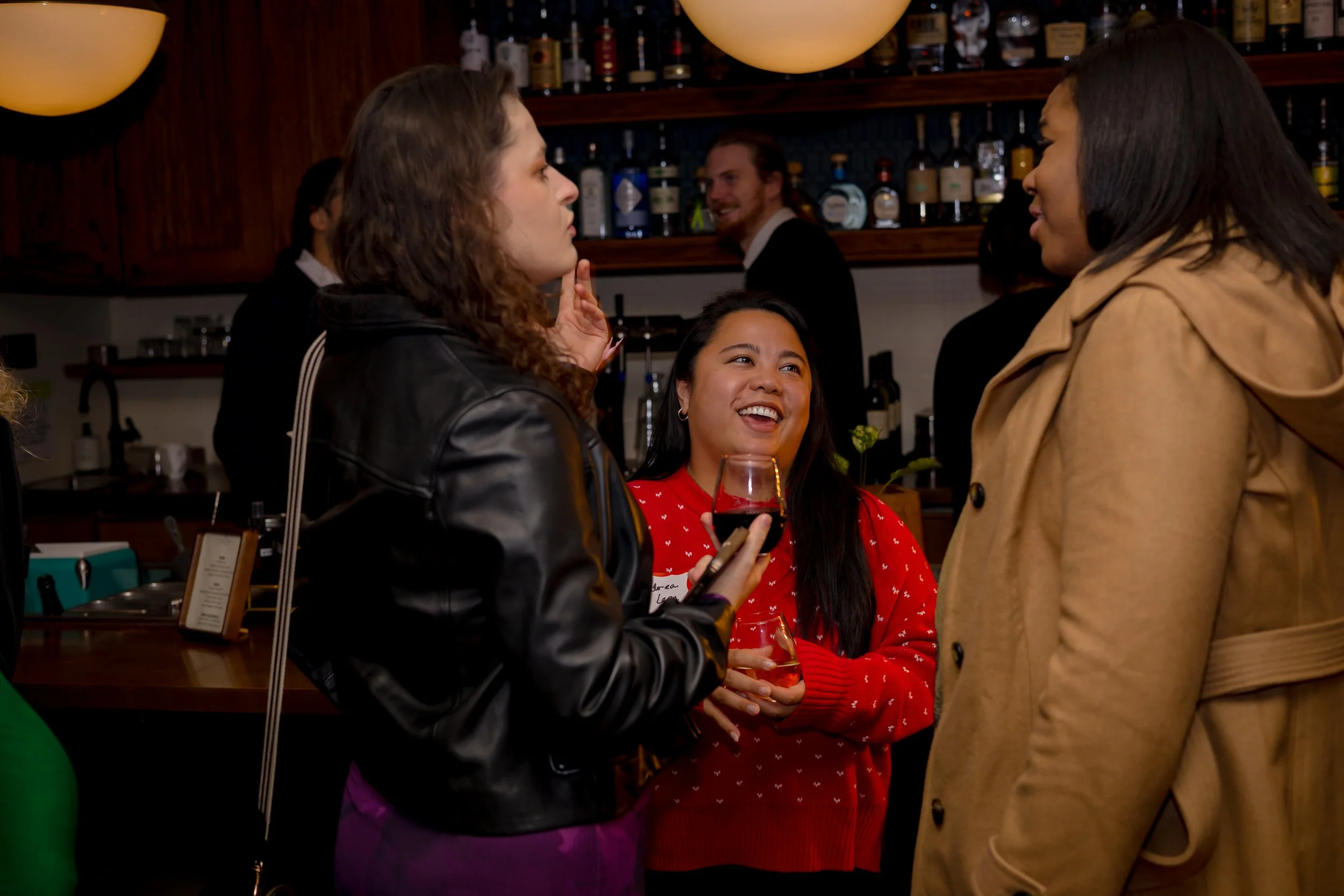 Three women are engaged in conversation at a bar or restaurant. The woman on the left is wearing a black leather jacket, the woman in the middle is wearing a red sweater with white patterns and holding a glass of red wine, and the woman on the right is wearing a tan coat. In the background, a bartender and another man are visible.