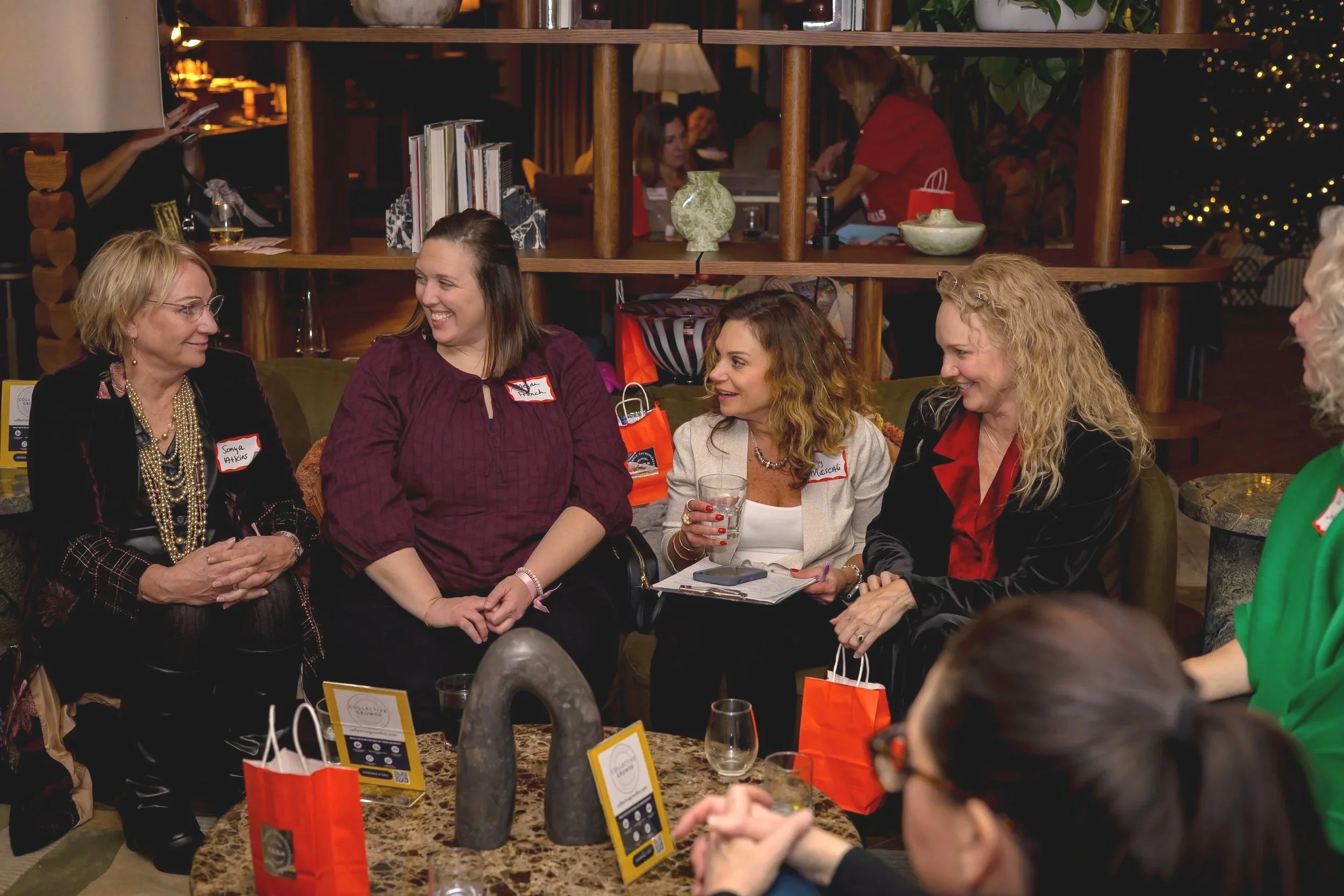 Group of women sitting together at a social gathering, talking and smiling. Some women wear name tags, and there are gift bags and drinks on the table in a warmly lit indoor setting decorated for the holidays.