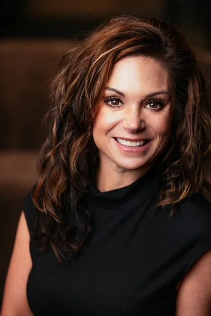 Founder Amy Mascali, a woman with wavy brown hair, smiles whiel wearing a black top, in a softly lit indoor setting.