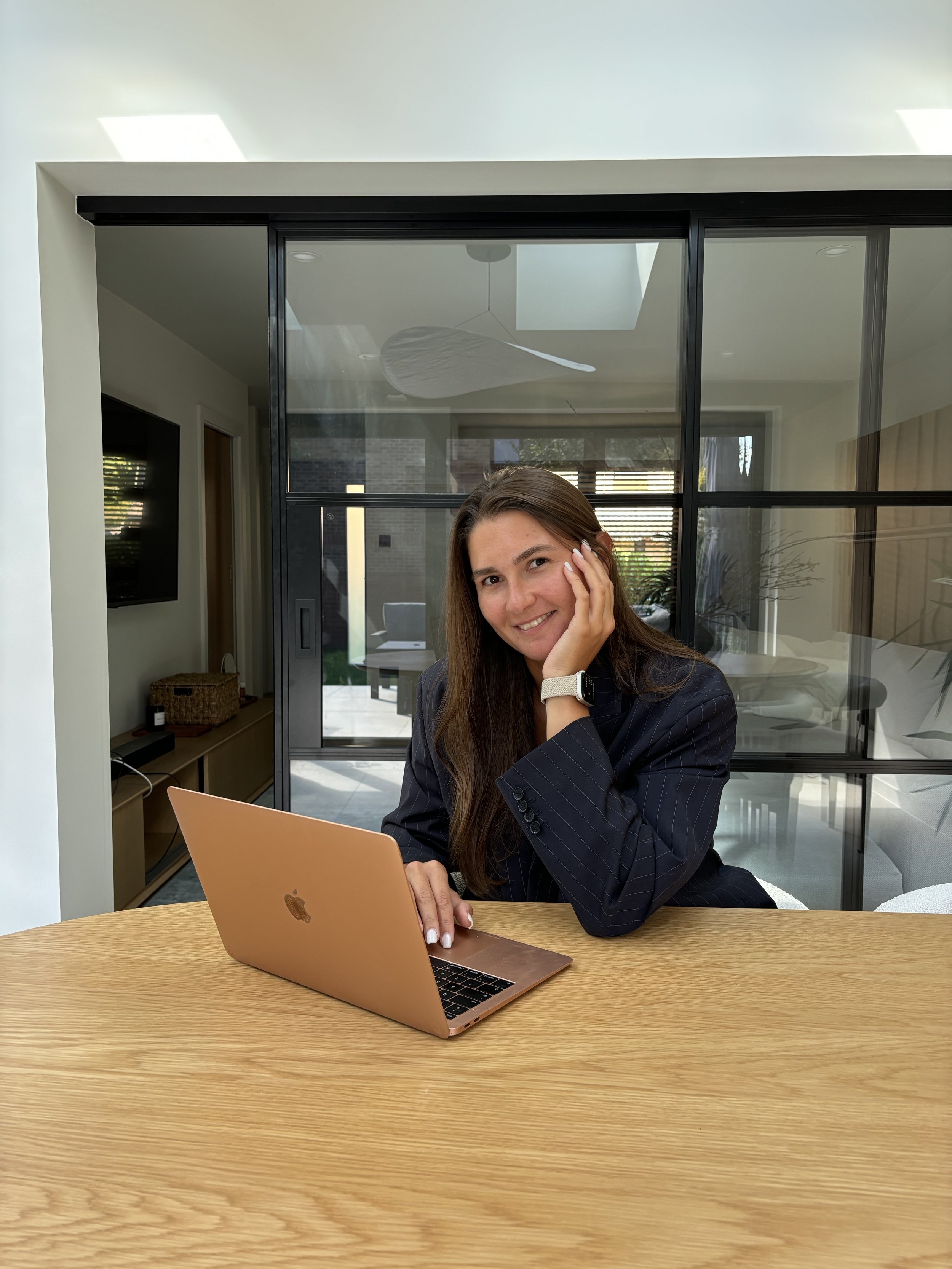 A woman with long brown hair smiling and resting her face on her left hand while sitting at a wooden table with a rose gold laptop in front of her. She is in a modern room with glass sliding doors and outdoor patio visible behind her.