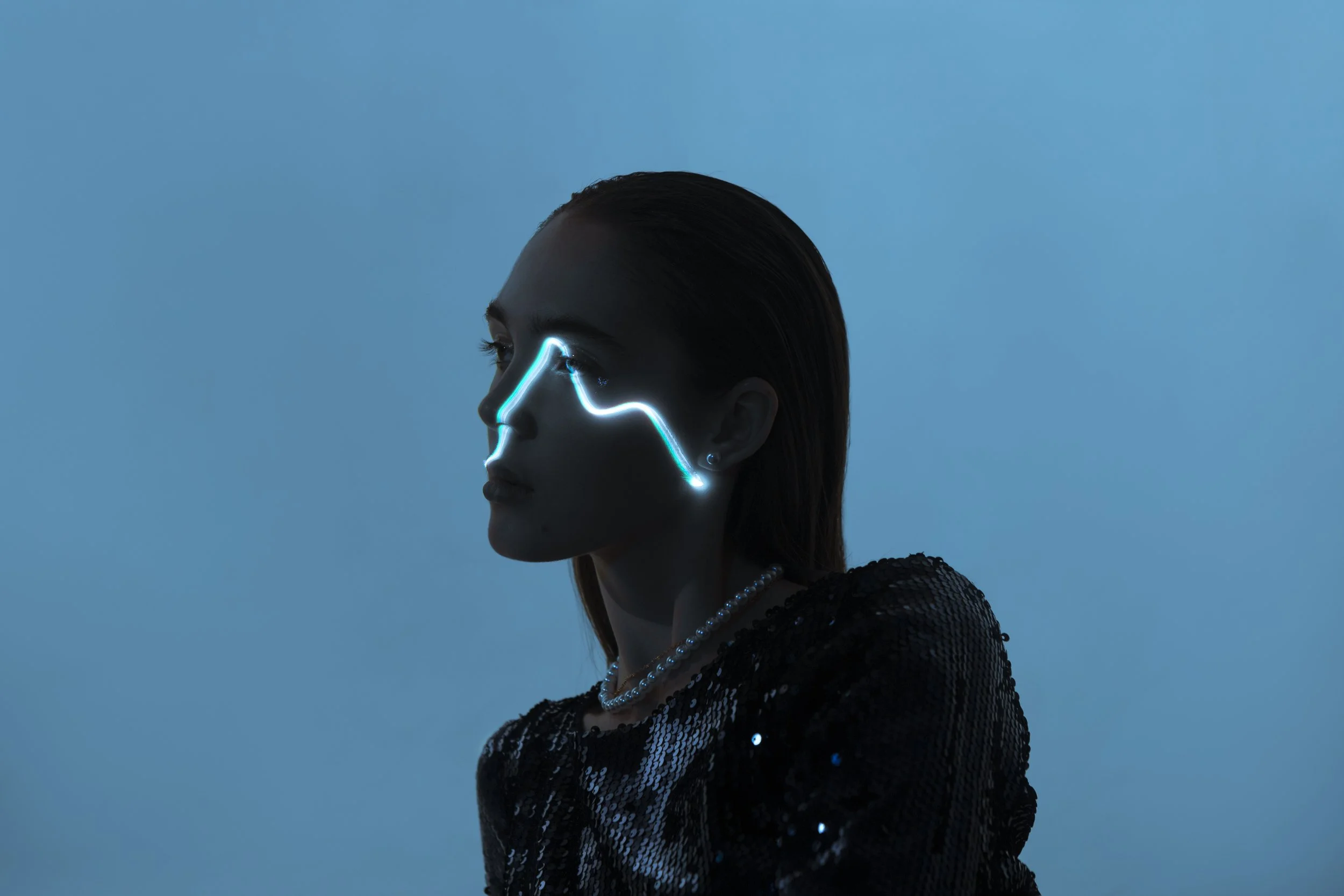 Side profile of a woman with neon blue lines on her face, wearing a black sequined top and pearl necklace, against a blue background.