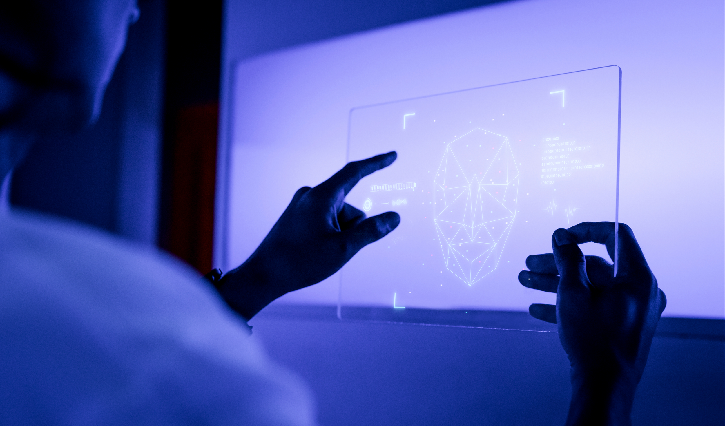 A person interacts with a futuristic computer display showing a digital wireframe face and scanning data.