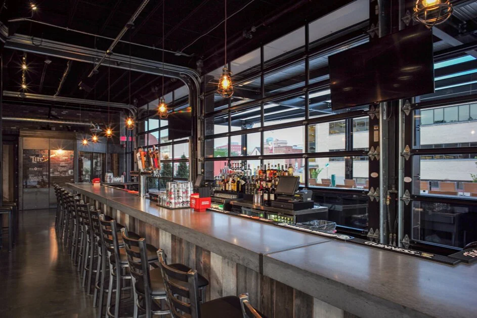 Empty bar with high stools, liquor bottles, beer taps, and large windows in a modern restaurant or bar.