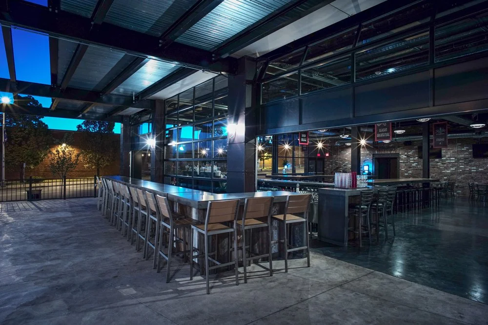 Empty indoor bar with bar stools, high-top tables, large windows, and a brick wall, illuminated by indoor and outdoor lighting during evening or night.