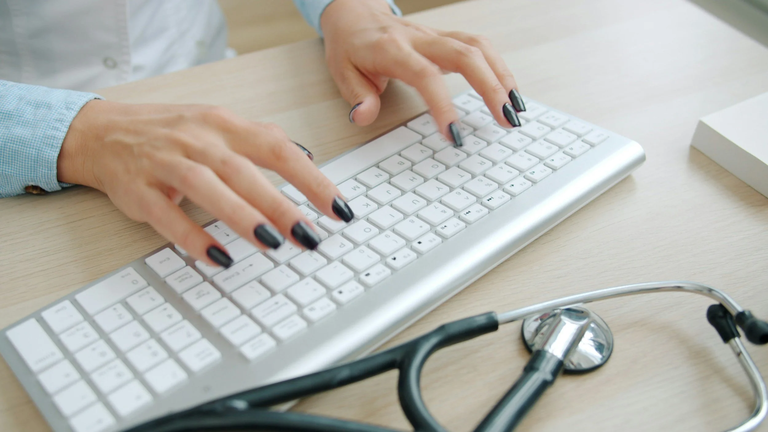 A person typing on a white keyboard with black painted nails, on a light wood desk with a stethoscope nearby.