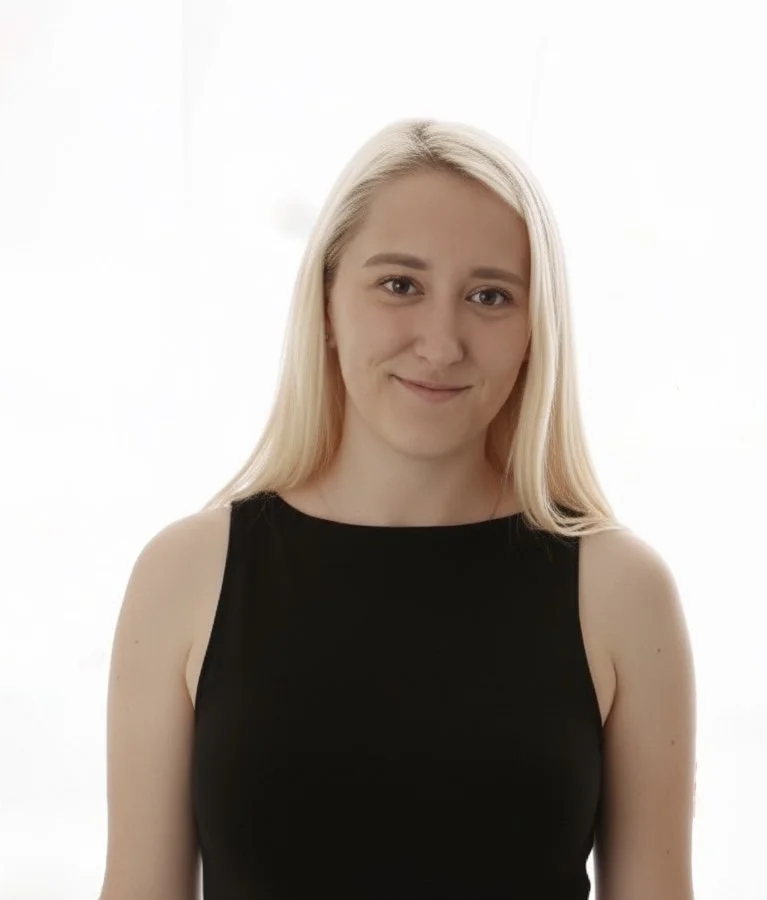 A young woman with long blonde hair wearing a black sleeveless top standing indoors against a white background.
