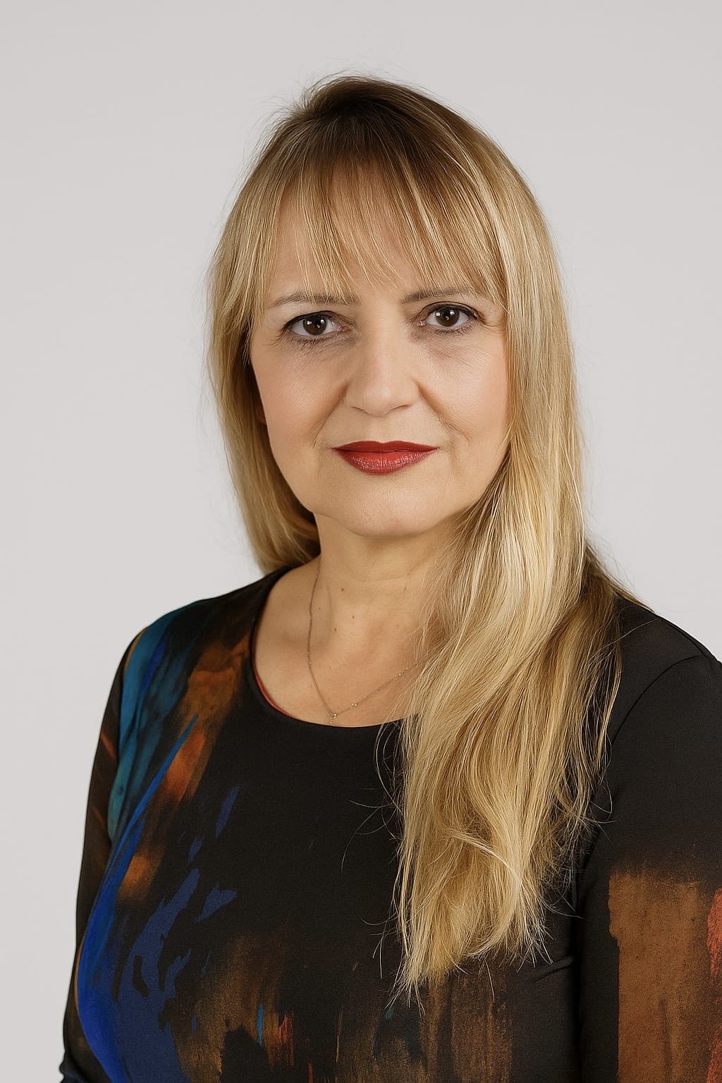 A woman with long blonde hair, wearing red lipstick, a black top with a colorful abstract pattern on one sleeve, and a gold necklace, standing against a plain white background.