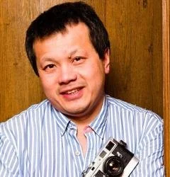 Mr Chan. Portrait of a man with short black hair wearing a blue and white striped shirt, standing against a wooden background, holding a vintage camera.