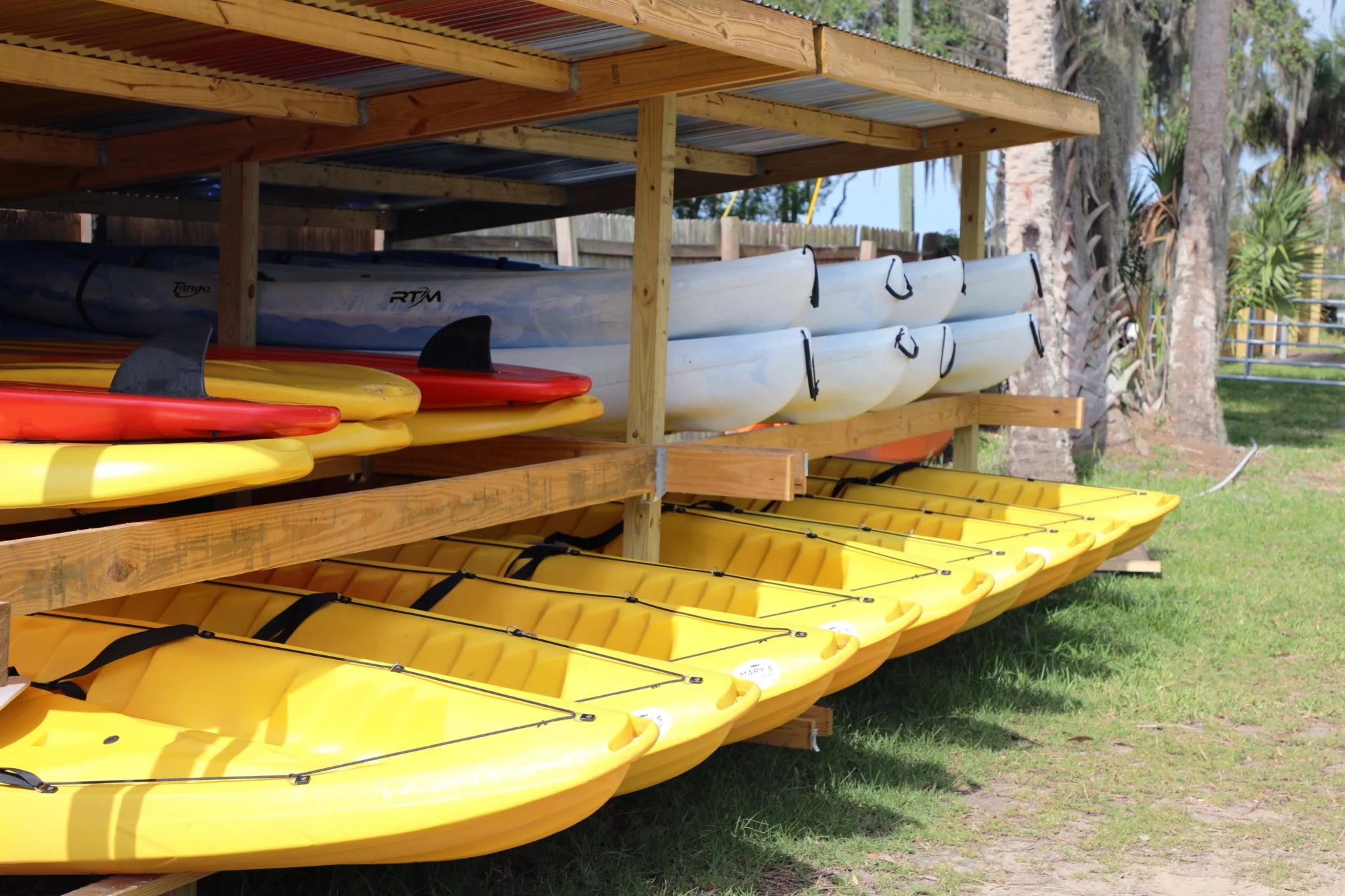 Stacked kayaks and paddleboards on a wooden rack with trees and grass in the background.