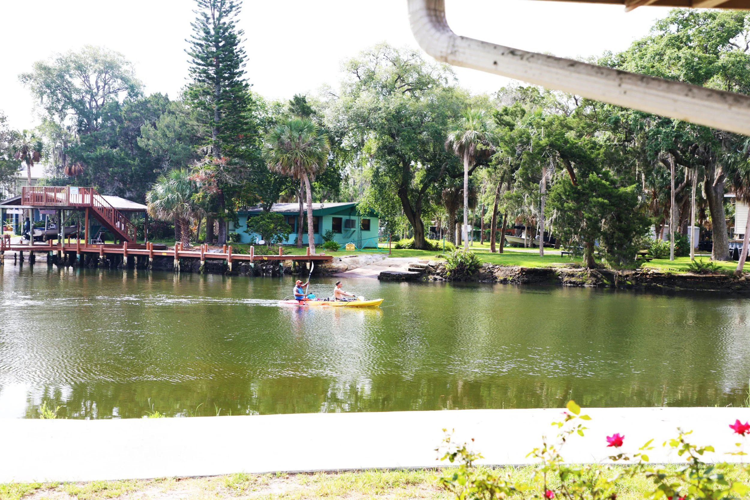 Two people kayaking in a yellow kayak on a calm river with lush green trees and houses in the background.