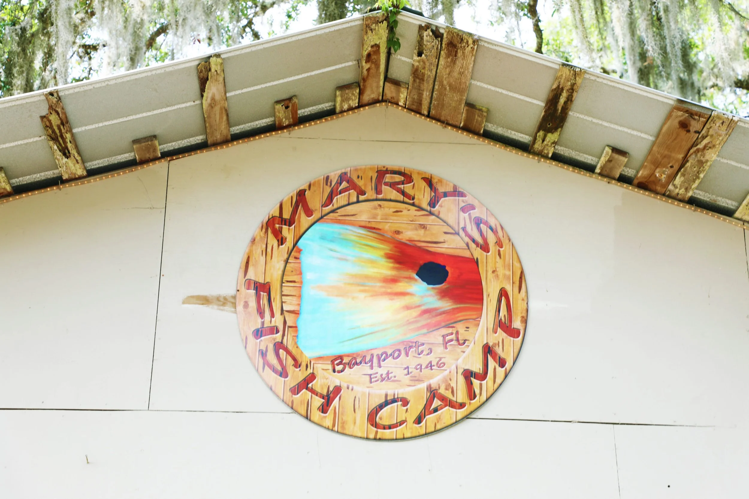 A circular sign that reads 'TIMARIS BEACH CAMPGROUND,' with a sunset scene depicting the sun setting over the water at Bayport, Florida, established in 1946, hung on the exterior wall of a building with a wooden roof and surrounded by trees.