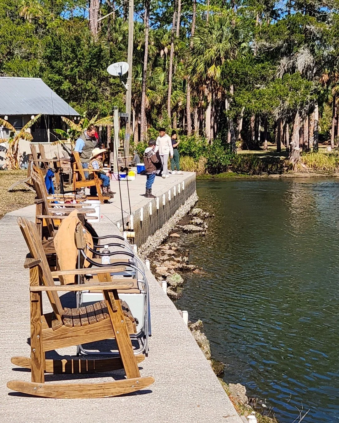 People fishing from a concrete dock by a lake, with wooden chairs lined up along the shore and lush green trees in the background.