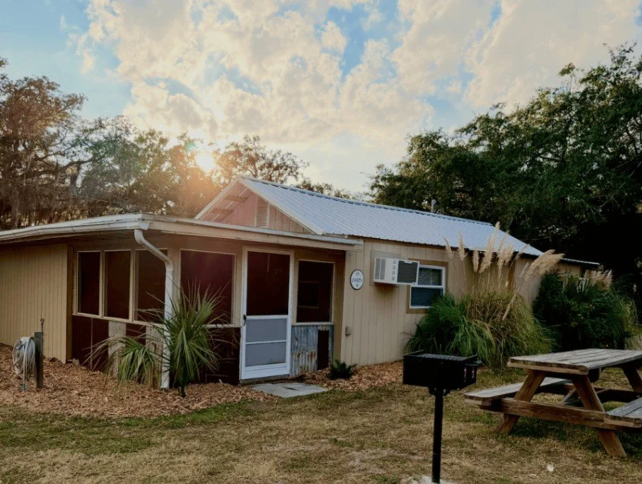 A suburban house with beige siding, a metal roof, an outdoor air conditioning unit, a screened porch, and a picnic table in the yard, with trees and a partly cloudy sky in the background.
