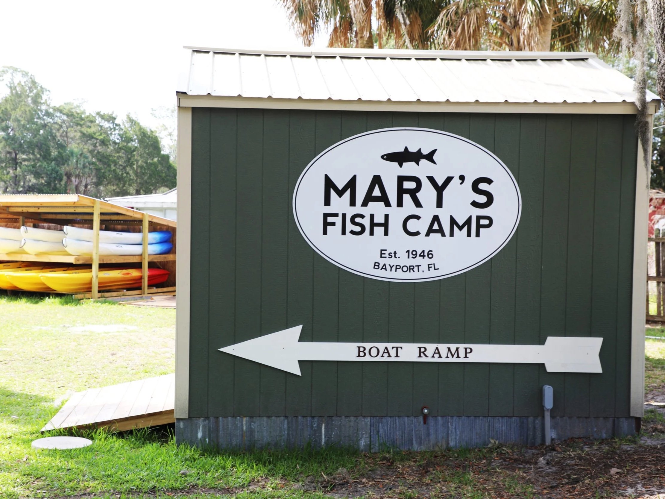 Sign for Mary's Fish Camp in Bayport, FL, with an arrow pointing left to the boat ramp, and a stack of kayaks in the background.
