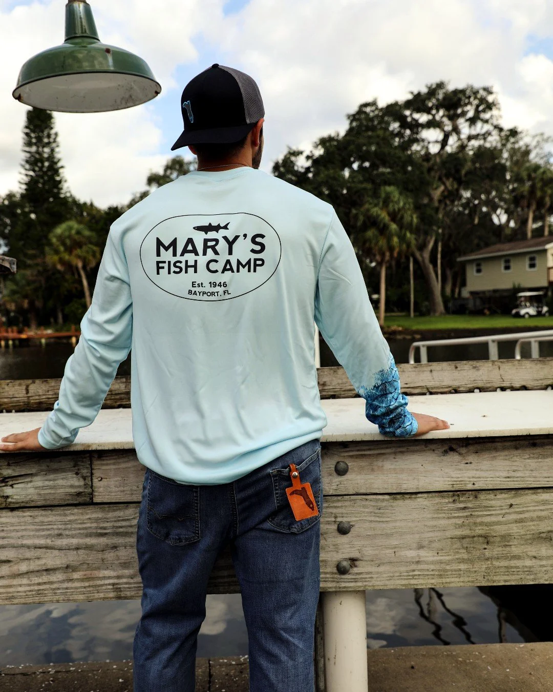 A man with his back to the camera, wearing a light blue long-sleeve shirt with 'Mary's Fish Camp' logo on the back, standing by a wooden railing overlooking a body of water with trees and houses in the background.