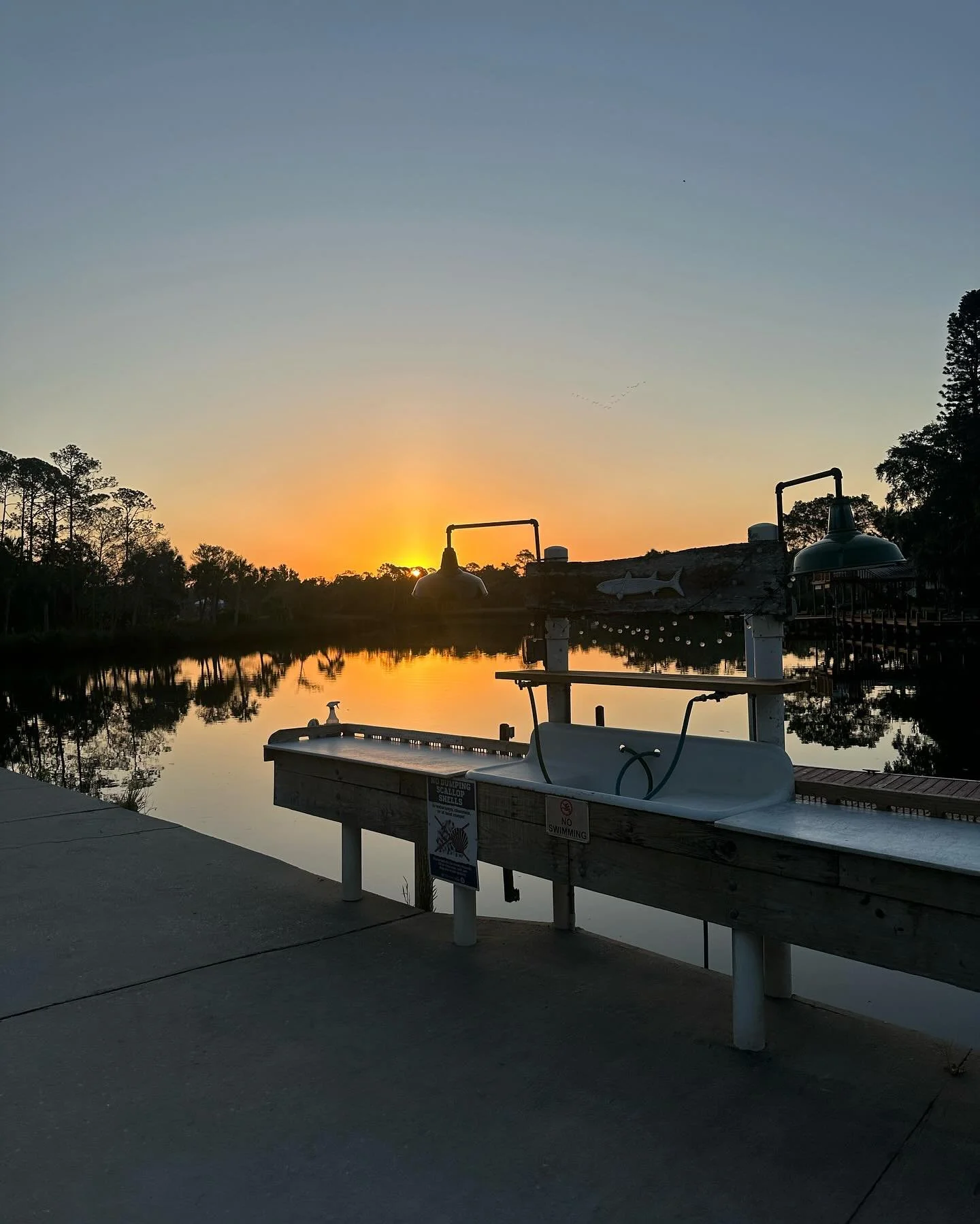 Sunset over a calm lake with silhouettes of trees and fishing equipment in the foreground.