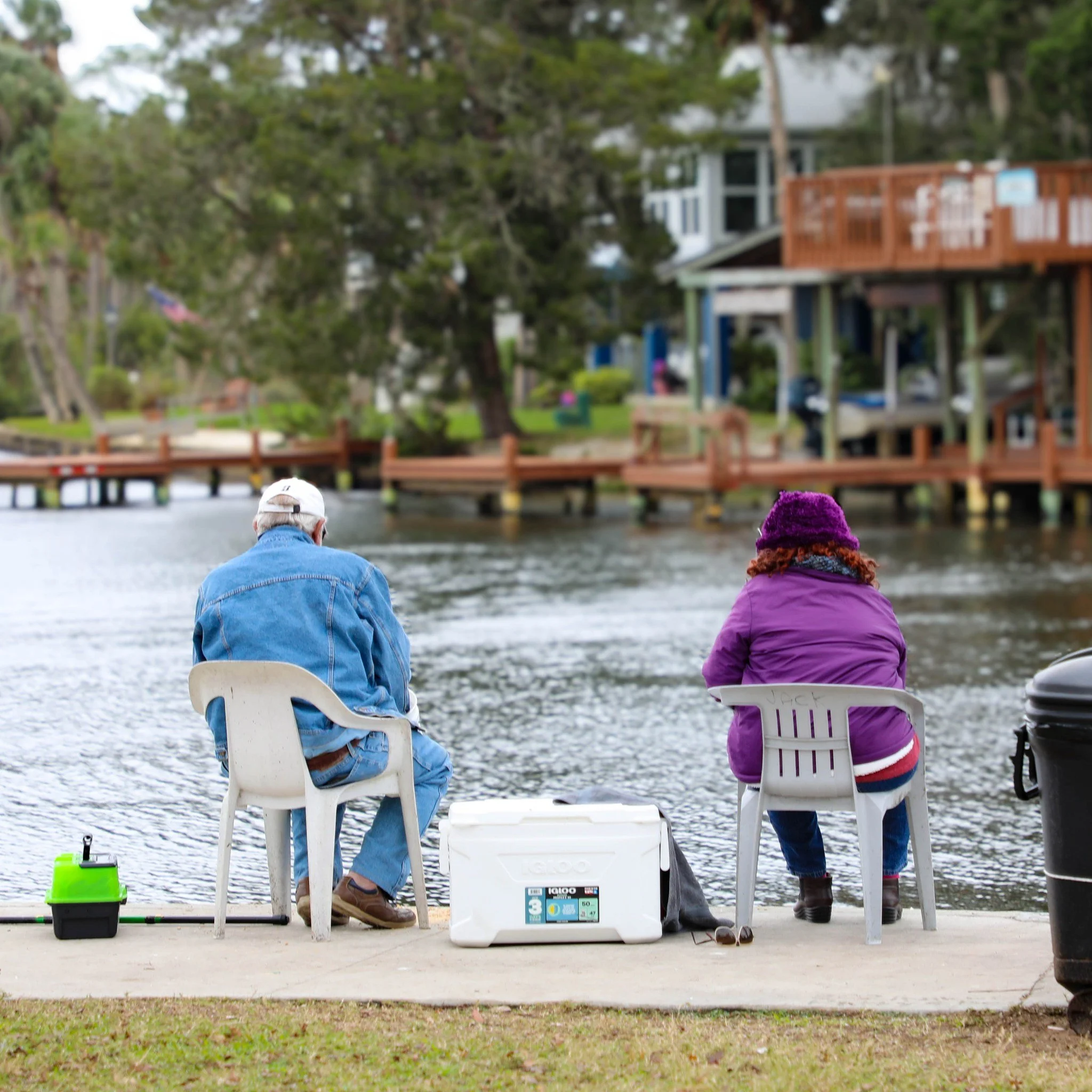 Two people fishing by a lakeside on a concrete dock, with houses and trees in the background.
