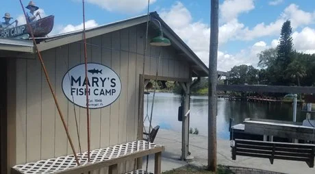 A fishing hut named Mary's Fish Camp located by a water body, with fishing nets hanging outside and boats visible in the background.