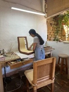A woman with long dark hair in a ponytail, wearing a white shirt and blue jeans, standing at a wooden desk with a cushioned chair in a cozy, rustic room with brick and white walls, a window with plants, and a ceiling light. Studio Luz, Barichara.