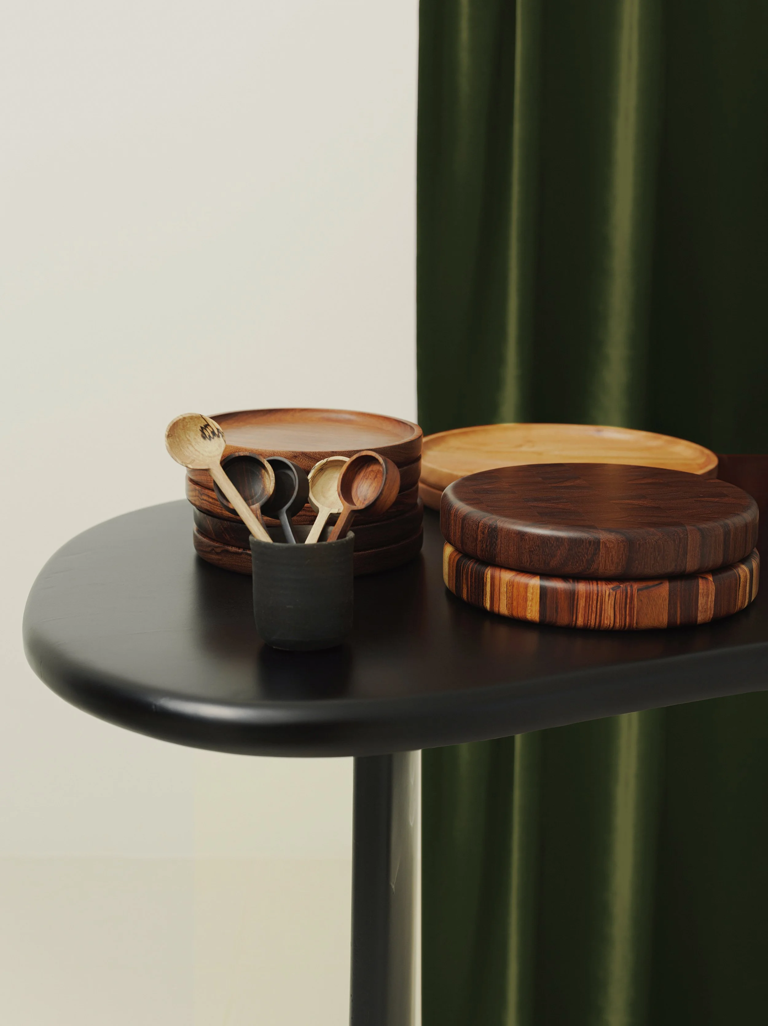Wooden plates in various shades stacked on a black table, with a black cup holding small wooden spoons, against a white wall and dark green curtains. Handcrafted by Itza Wood, Guatemala.