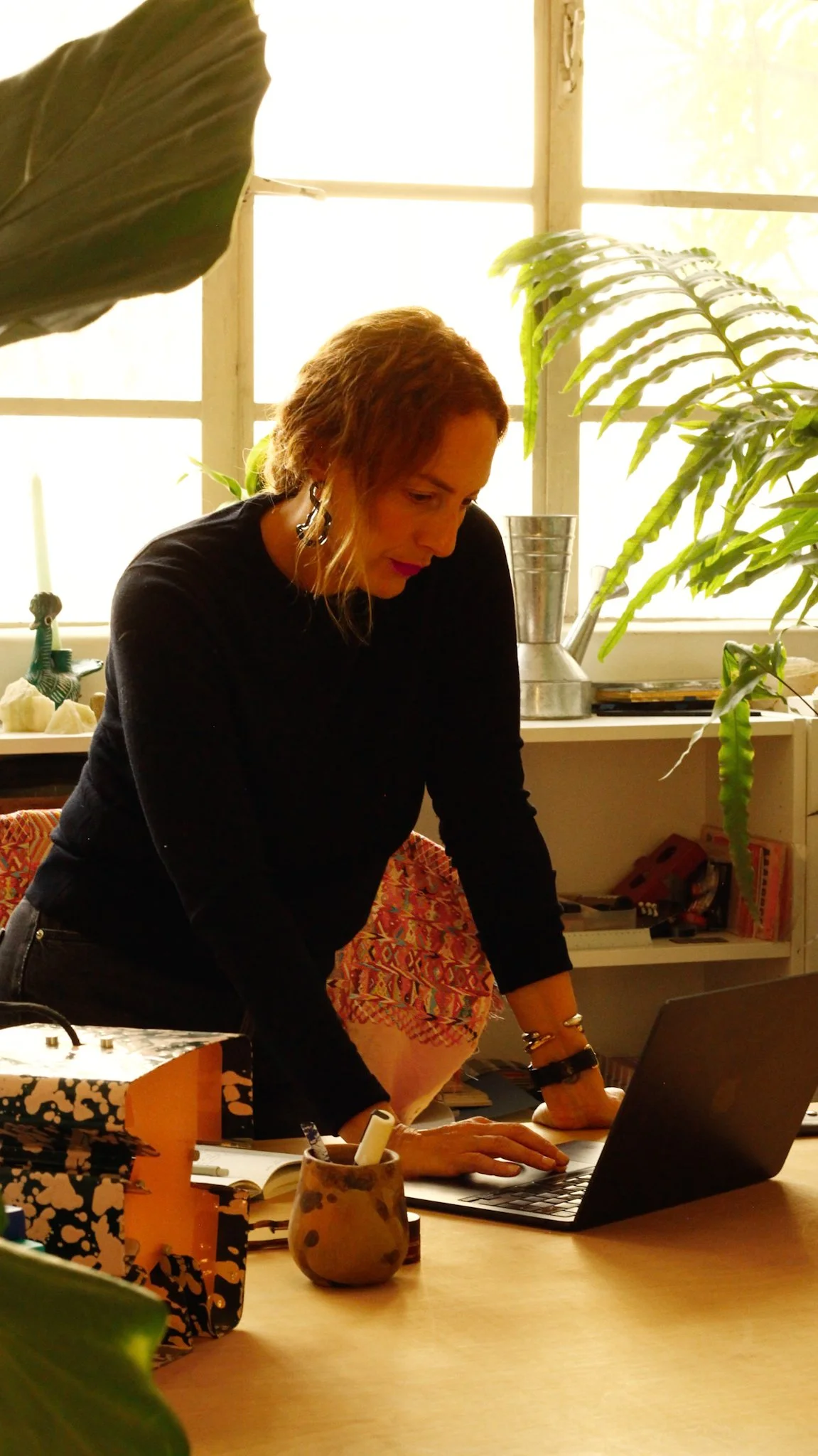 A woman working at a desk with a laptop in a bright room filled with large green plants and sunlight.