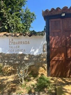 A white wall with a sign that reads 'La Tabarrada Taller Cerámica' next to a wooden gate, with green trees and a clear blue sky in the background. Shot in Barichara, Colombia.