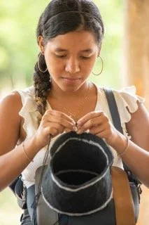 Young woman with braided hair and hoop earrings, looking down and holding a necklace or bead string. She carries a bag with a sport shirt printed on it. In Atanquez, Colombia.