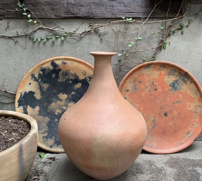 Four pottery pieces, including a large, rounded clay vase and three flat, round trays with weathered surfaces, arranged outdoors against a concrete wall with vine growth. Made in Mexico.