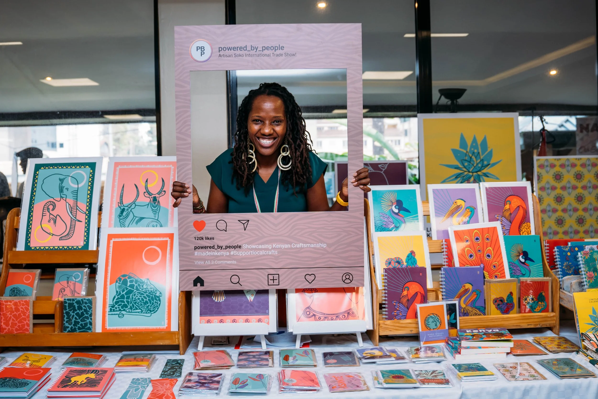 A woman smiling and holding a large Instagram frame cutout at Artisan Soko Nairobi, Kenya with various colorful paintings and artworks of animals and nature.