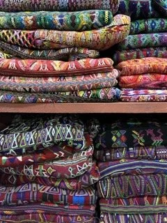 Stack of folded colorful huipiles with various geometric and floral patterns from Guatemala.