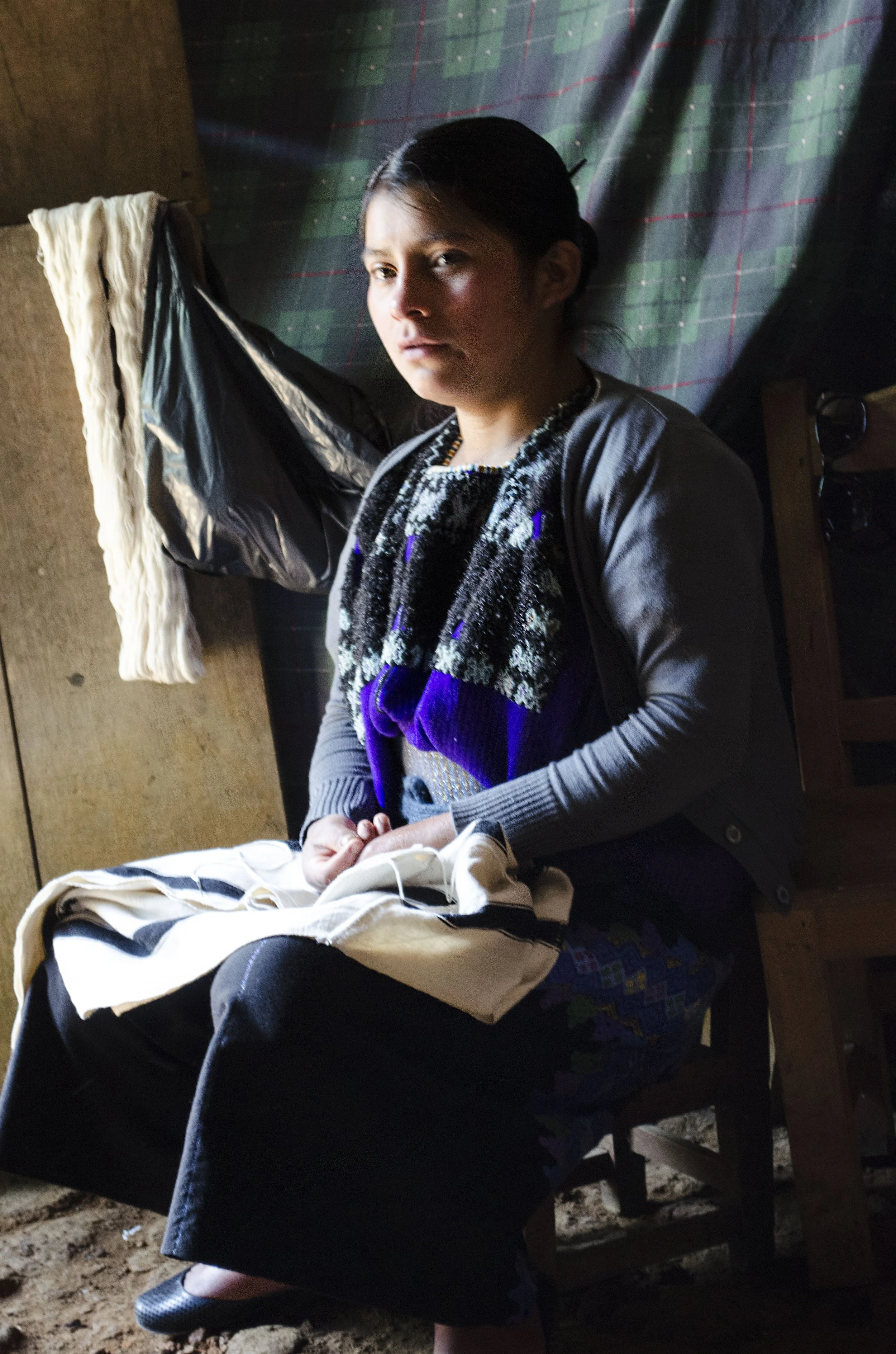 A weaver from Chiapas Mexico sitting on a wooden chair inside a dimly lit room, with colorful fabric hanging in the background, holding a cloth and looking at the camera.