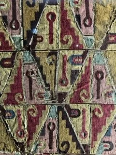 Close-up of a colorful, geometric patterned textile or artwork featuring red, yellow, and blue colors with key and staircase motifs. Made in Peru.