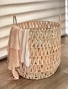 A round woven woven basket with two handles, placed on a wooden floor next to a window blind, with a light-colored scarf draped over its side.